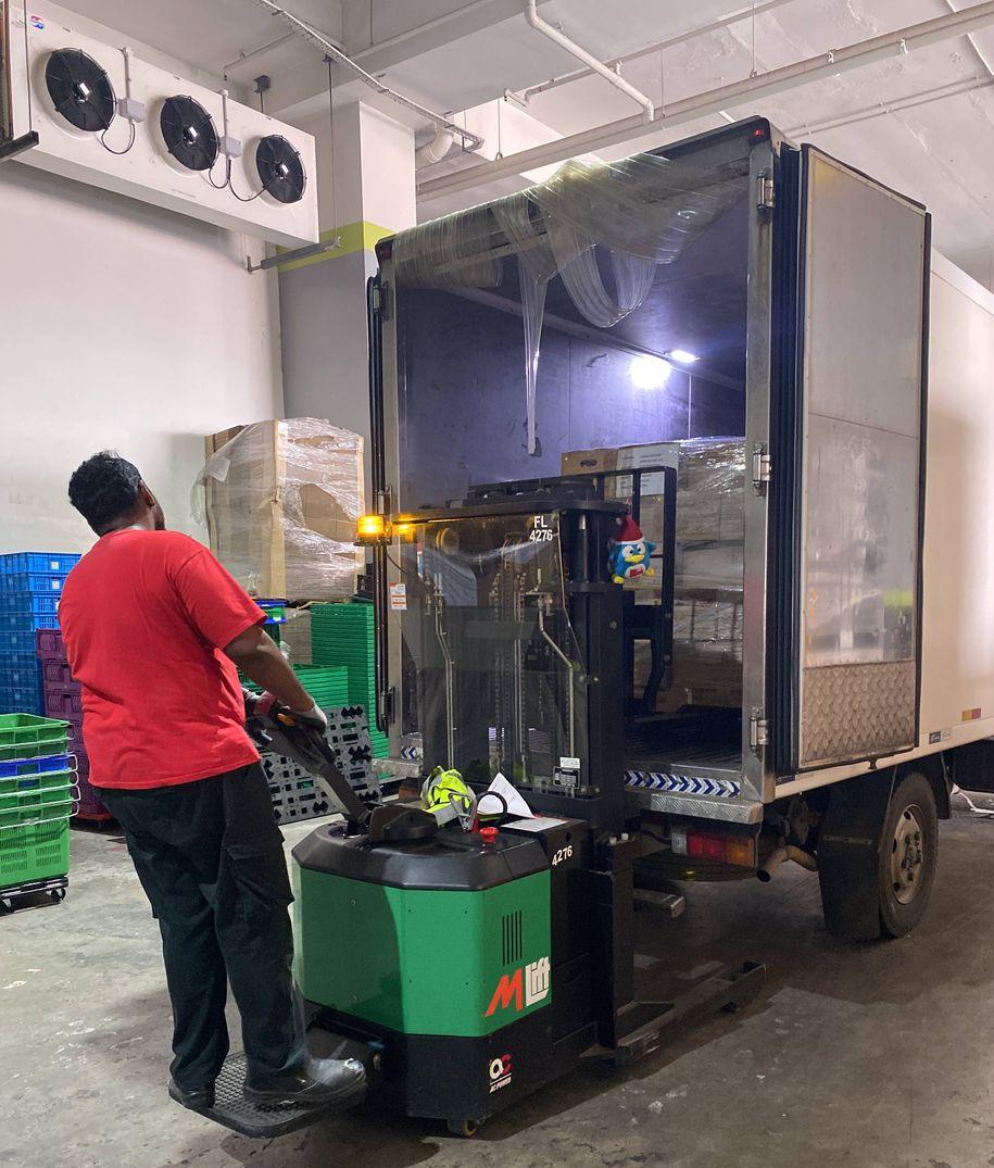 Worker using a green electric forklift to unload a refrigerated truck at a cold-room loading bay